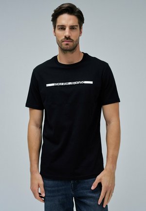 Hombre con cabello oscuro vistiendo una camiseta negra con el texto "SALSA JEANS" en el pecho y jeans azules, de pie contra un fondo gris liso.