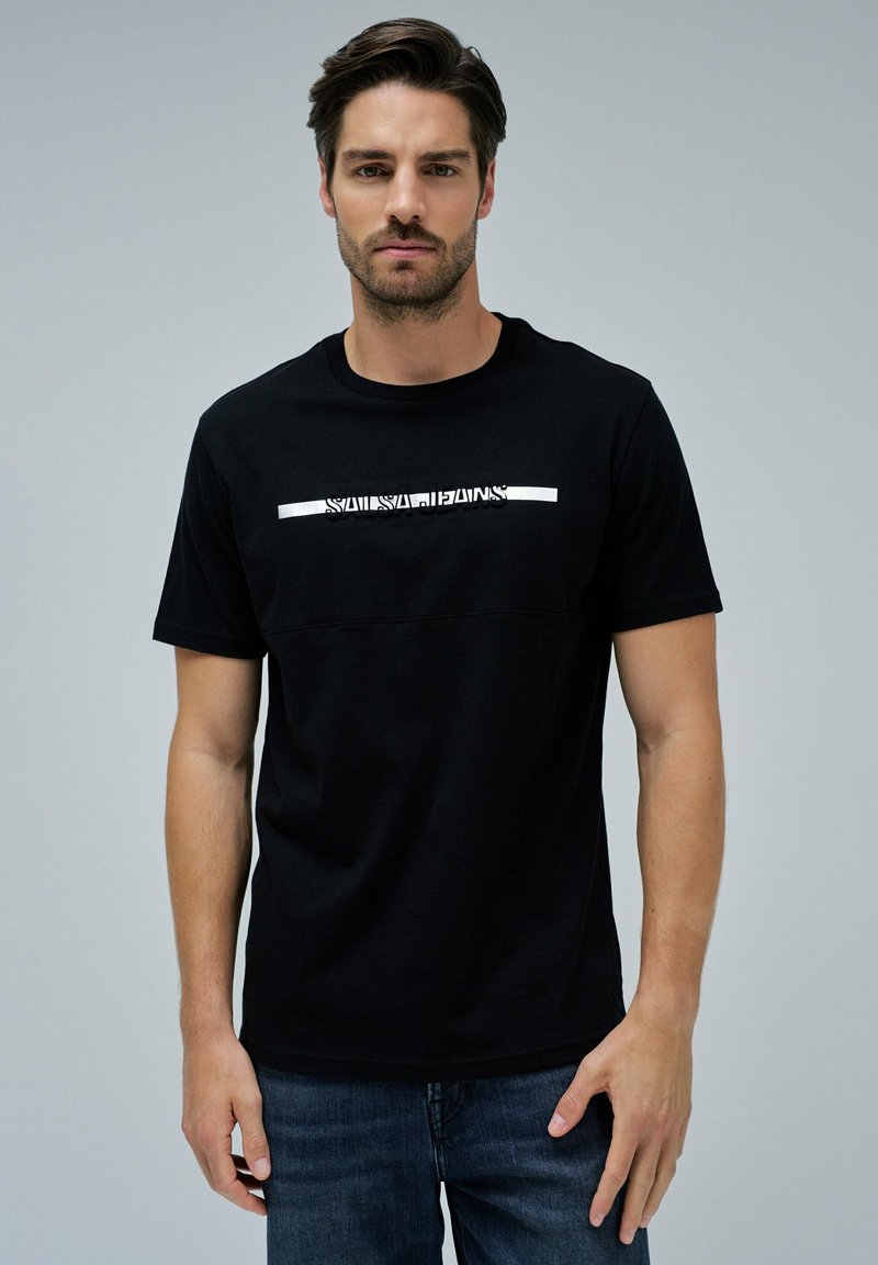 Hombre con cabello oscuro vistiendo una camiseta negra con el texto "SALSA JEANS" en el pecho y jeans azules, de pie contra un fondo gris liso.