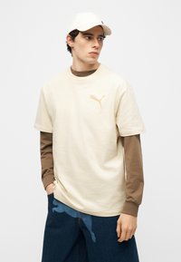 Νέος άνδρας που φοράει μπεζ μπλουζάκι Puma πάνω από καφέ πουλόβερ με μακριά μανίκια, μπλε τζιν και λευκό καπέλο, στέκεται με τα χέρια στις τσέπες.