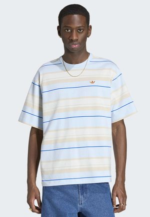 Młody mężczyzna w jasnoniebieskiej, beżowej i niebieskiej pasiastym t-shircie Adidas oraz niebieskich dżinsach, stojący na jednolitym tle.