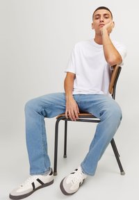 Hvit bomulls t-skjorte, lyseblå denimjeans og hvite sneakers med sorte detaljer. Sittende på en trestol med metallramme.