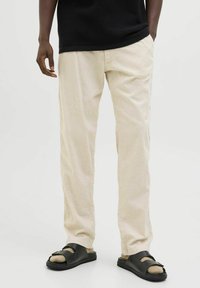 Béžové kalhoty s rovnými nohavicemi z lehké látky, doplněné černými nazouvacími sandály s přezkou a texturovanou podrážkou.