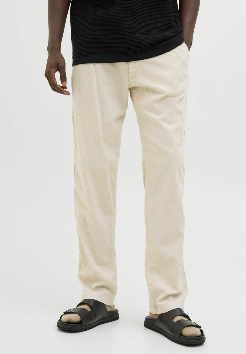 Béžové kalhoty s rovnými nohavicemi z lehké látky, doplněné černými nazouvacími sandály s přezkou a texturovanou podrážkou.