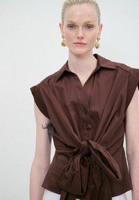 Camicia marrone a maniche corte con vita legata, dotata di colletto e chiusura a bottoni. Texture liscia con una vestibilità strutturata.