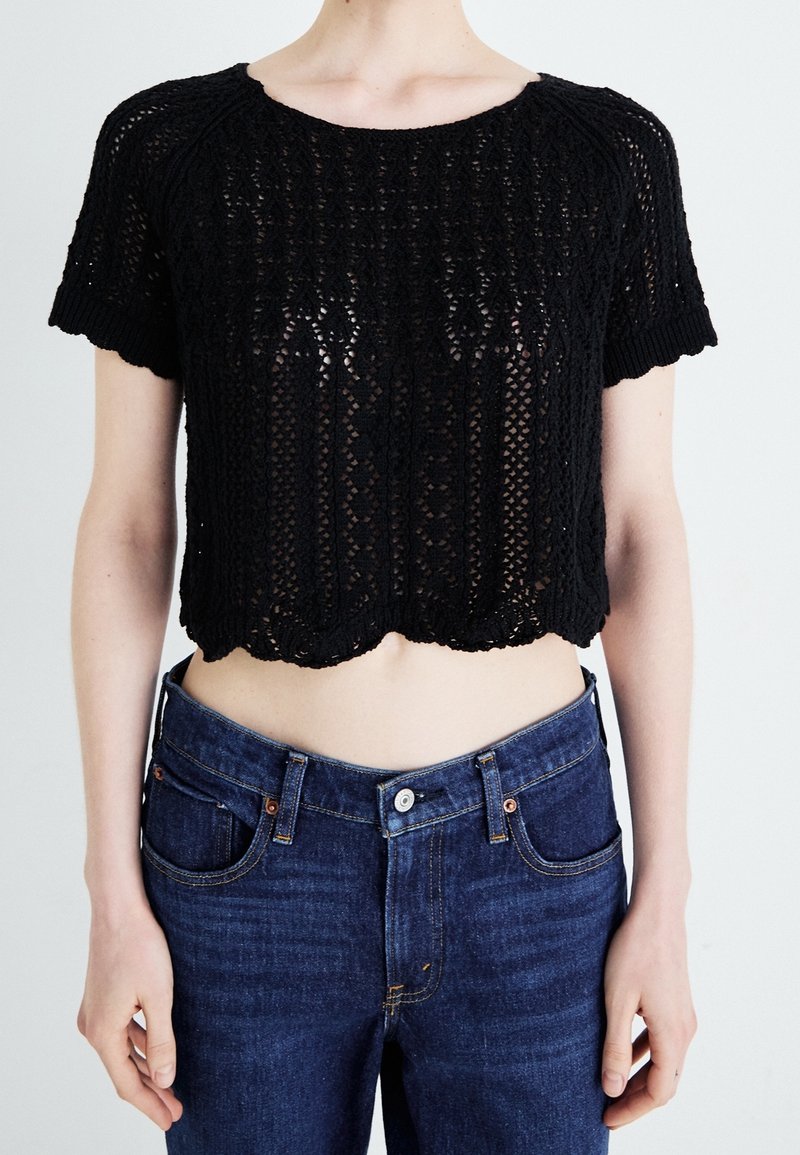 Personne portant un haut court en crochet noir à manches courtes et un jean bleu taille haute, debout devant un fond uni.