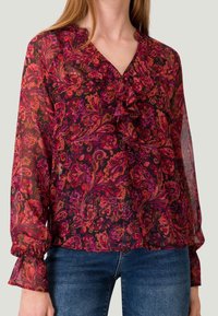 Blouse noire transparente avec des motifs floraux paisley vibrants en rose et orange, col froncé et poignets élastiques. Portée avec un jean bleu.