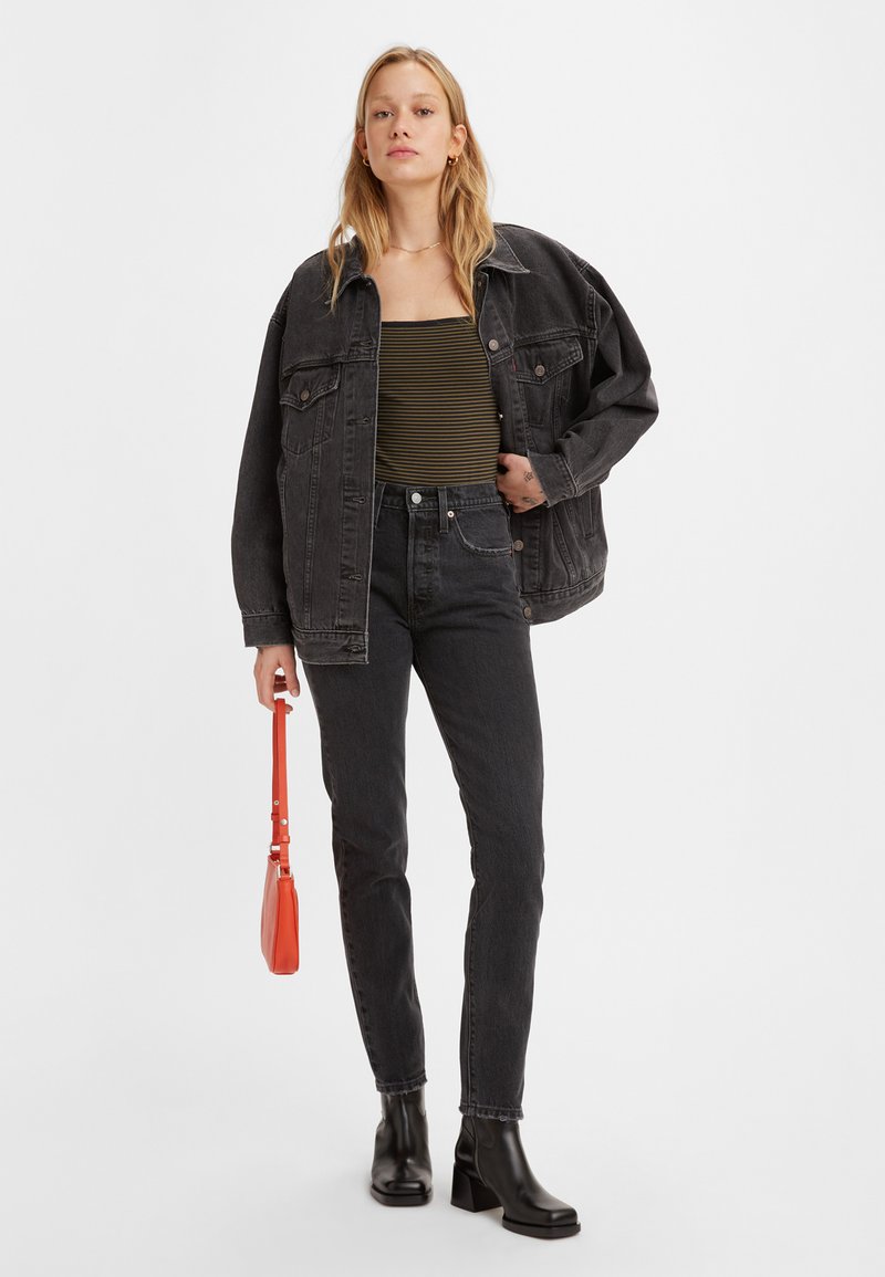 Schwarze Denimjacke, gestreiftes grünes Oberteil, schwarze High-Waist-Jeans und schwarze Stiefel mit Blockabsatz. Hält eine kleine rote Handtasche. Einfarbiger Hintergrund.