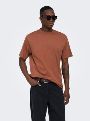 Mand iført brun t-shirt, sorte solbriller, sorte bukser, brun bælte, står med den ene hånd i lommen, tiger-tatovering på venstre underarm.