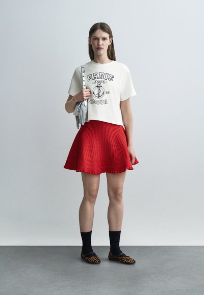 Νεαρή γυναίκα στέκεται φορώντας λευκό μπλουζάκι με τη φράση «Paris Amour», κόκκινη πλισέ φούστα, μαύρες κάλτσες, παπούτσια με λεοπάρ μοτίβο και ασημένια τσάντα ώμου με φούντες.