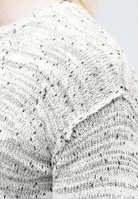 Stickad tröja i vitt med svarta prickar, med rund halsringning och korta ärmar. Tydligt strukturerad mönster och fransig nederkant.