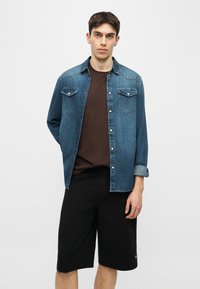 Jeune homme portant une chemise en denim bleue à boutons par-dessus un t-shirt marron et un short noir jusqu'aux genoux, debout devant un fond uni.