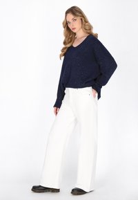 Marineblauer V-Ausschnitt-Strickpullover mit Glitzer, kombiniert mit weißen weit geschnittenen Hosen und schwarzen Schuhen mit dickem Absatz. Glatte Textur und lockere Passform.