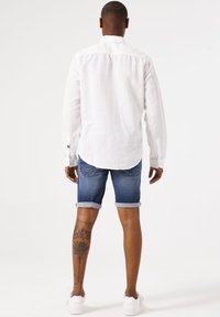 Camisa blanca de manga larga con puños de botón, combinada con shorts de mezclilla enrollados. Zapatillas blancas, tatuaje floral visible en la pantorrilla. Fondo neutro.
