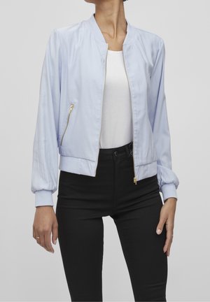 Femme portant une veste bomber bleu clair par-dessus un haut blanc et un pantalon noir, debout avec les bras détendus le long du corps devant un fond uni.