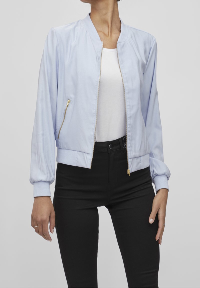 Femme portant une veste bomber bleu clair par-dessus un haut blanc et un pantalon noir, debout avec les bras détendus le long du corps devant un fond uni.
