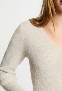 Mujer vestida con un suéter de punto suave y ligero en color beige claro, con escote en V, visible parcialmente desde el hombro hasta la parte superior del pecho.