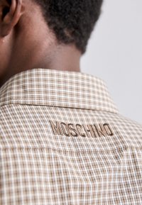 Kariertes braunes und beiges Hemd mit steifem Kragen, auf dessen Rückseite "MOSCHINO" in braunem Faden gestickt ist. Weicher Stoff.
