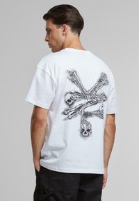 Hvid bomulds T-shirt med en sort grafik af to kranier og korslagte knogler på ryggen. Kortærmet og afslappet pasform.