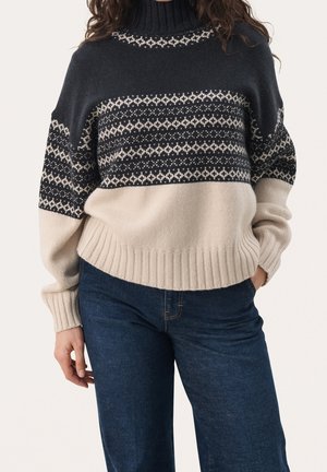 Frau trägt einen hochgeschlossenen schwarzen und beigen Pullover mit Muster, mit gerippten Bündchen und Saum, kombiniert mit dunkelblauen Jeans, Hand in der Tasche.