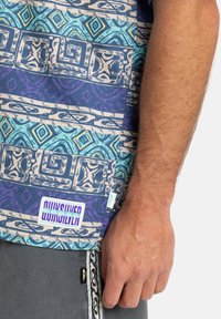 Camisa de manga corta con tela estampada en tonos de turquesa, azul y púrpura, con una etiqueta de logo en el bajo y diseños texturizados.