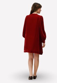 Rode fluwelen jurk met een losse pasvorm, lange pofmouwen en een recht afgesneden zoom. Soepele textuur en minimale details.