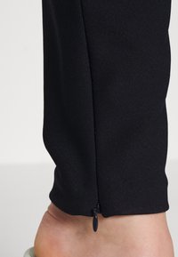 Svart tapered byxben med en slät textur, med en liten dragkedjedetalj vid fållen. En bar fot syns vid botten.