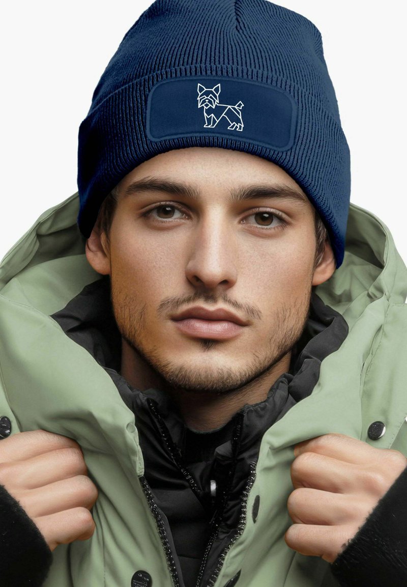 Navy knit beanie featuring a textured pattern and a rectangular patch with a white dog graphic, worn with a green puffer jacket.