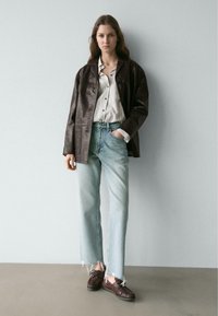 Dunkelbraune Lederjacke über einem gestreiften Hemd mit Knöpfen, kombiniert mit hellblauen Jeans mit ausgefranstem Saum und dunkelbraunen Schuhen. Schlichter Hintergrund.