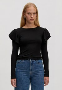 Black ribbed long-sleeve top with ruffled shoulder accents, paired with high-waisted blue denim jeans. Smooth texture, round neckline.