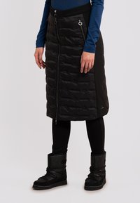 Falda acolchada negra con un patrón de acolchado horizontal, que presenta bolsillos laterales con cremallera y un diseño dividido. Combinada con botas de invierno negras y brillantes.