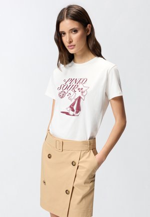 Femme portant un t-shirt graphique blanc avec le texte « PINKO SOUR » et un personnage étoile, associé à une jupe beige ceinturée avec des détails de boutons.