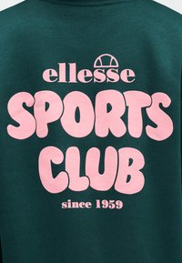 Σκούρο πράσινο ύφασμα με μεγάλο ροζ κείμενο "SPORTS CLUB" και μικρότερο λογότυπο "ellesse" και το κείμενο "since 1959" τυπωμένα πάνω του.