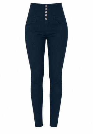 Vrouw die hoog getailleerde donkerblauwe skinny jeans draagt met vijf zilveren knopen en witte platform sneakers, staand tegen een effen achtergrond.