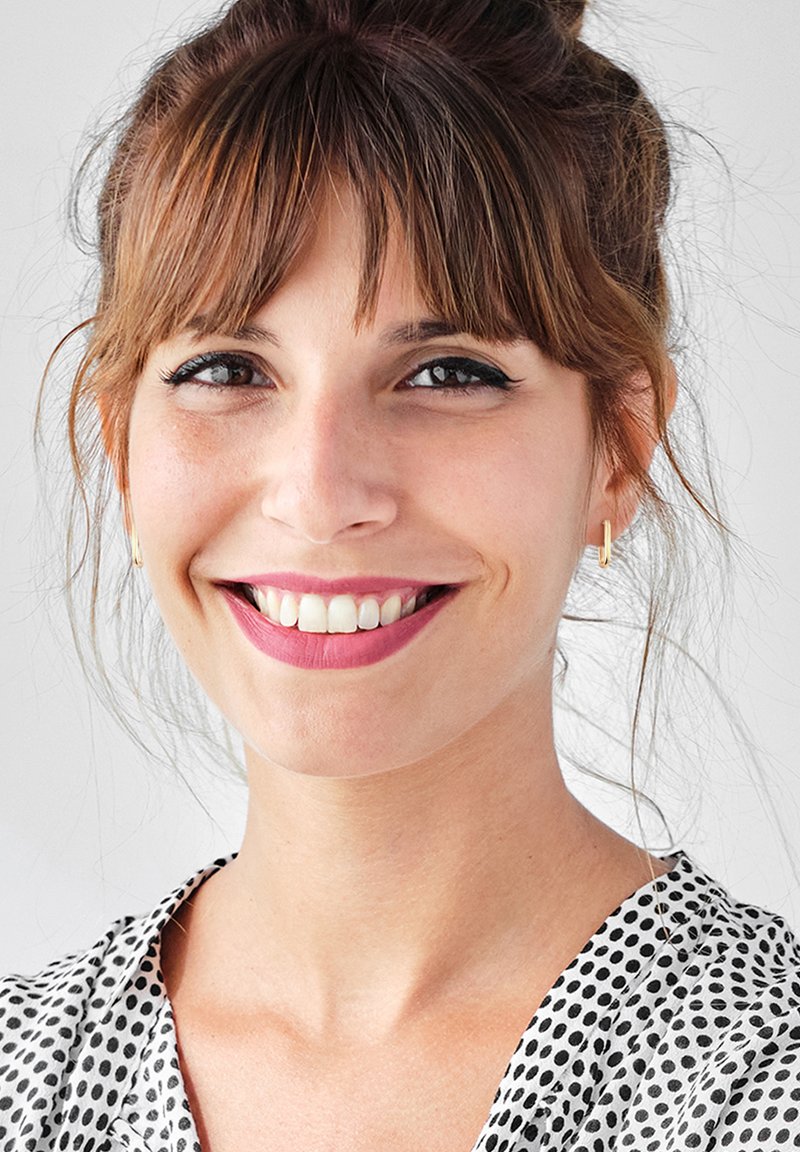 Femme souriante avec des cheveux bruns coiffés en chignon, portant de petites boucles d'oreilles en or, et une chemise à pois noir et blanc. Fond lumineux.