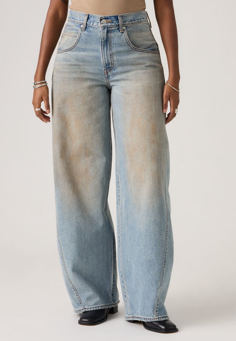 Άτομο που φοράει ανοιχτού μπλε χρώματος τζιν παντελόνι με φαρδιά μπατζάκια, μπεζ μπλούζα, μαύρα παπούτσια με τακούνι και ασημένια βραχιόλια και δαχτυλίδια και στα δύο χέρια.