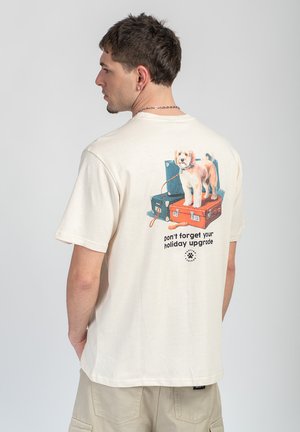 Man wearing cream T-shirt with graphic of dog standing on stacked blue and orange suitcases and text "don’t forget your holiday upgrade."