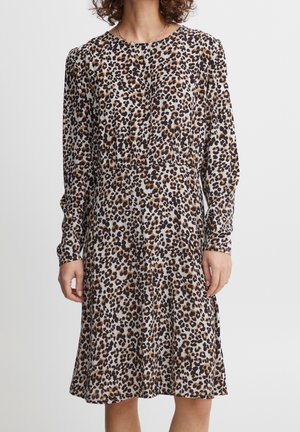 Kobieta ubrana w sukienkę sięgającą do kolan z długim rękawem, z brązowo-czarnym wzorem w cętki na jasnym tle.