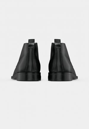 Vue arrière d’une paire de bottines en cuir noir avec des tirettes et des semelles épaisses et plates, sur un fond clair uni.