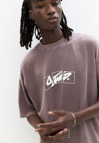 T-shirt oversized viola con un logo grafico bianco sulla parte anteriore. Realizzato in cotone morbido, con scollatura rotonda e maniche corte.
