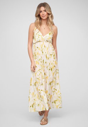 Femme aux longs cheveux ondulés portant une robe longue fleurie jaune et blanche, des sandales et un collier, debout devant un fond uni.