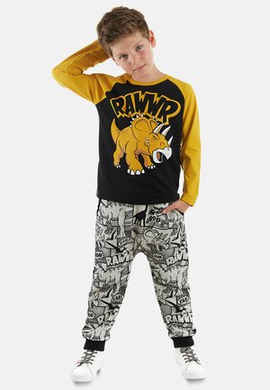 Schwarz-gelbes Langarmshirt mit einem Dinosaurier-Graphic; graue gemusterte Jogginghose mit Dinosaurier- und Geräuscheffekten; weiße Sneakers.