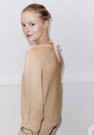 Beige Strickpullover mit geripptem Muster und 3/4-Ärmeln, mit einer kleinen satinierten Schleife am oberen Rücken. Heller Hintergrund.