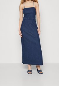 Robe maxi en denim bleu foncé avec des bretelles fines, un buste ajusté et une jupe évasée. Associée à des chaussures à talons hauts navy avec un détail de nœud.