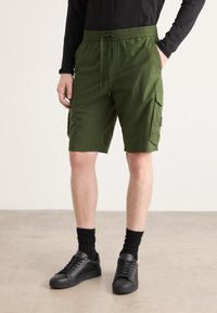 Grønne cargo shorts har et elastisk liv med snøring, sidelommer og en glatt tekstur. Kombinert med svarte sneakers.