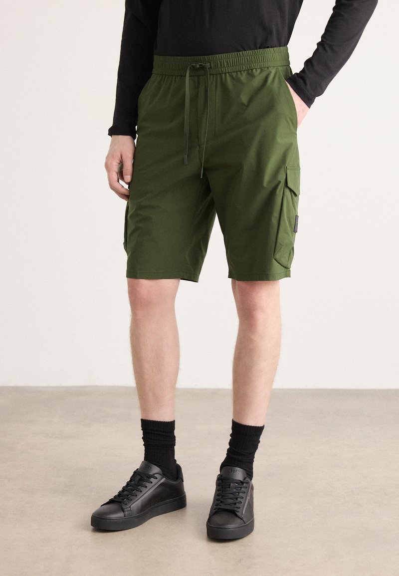 Grønne cargo shorts har et elastisk liv med snøring, sidelommer og en glatt tekstur. Kombinert med svarte sneakers.