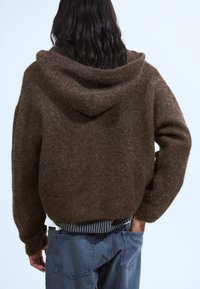 Un sweat à capuche marron en tissu doux et texturé. Il présente une coupe ample et un ourlet inférieur côtelé, associé à un jean bleu décontracté.