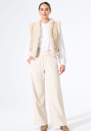 Vrouw in een beige faux fur-geborduurde vest, witte blouse met lange mouwen, brede crèmekleurige broek en bruine loafers, staand met één hand in de zak.