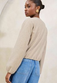 Beige, struktureret sweater med ballonærmer og en samlet kant, parret med lyseblå denim jeans med baglommer. Guld øreringe synlige.