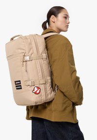 Beige ryggsäck med justerbara remmar, flera fack och en 'Ghostbusters'-logotypapplikation. Materialet är slitstark tyg; har spänneslås.