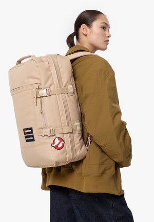 Mochila beige con correas ajustables, múltiples compartimentos y un parche del logo de 'Cazafantasmas'. El material es de tela resistente; cuenta con cierres de hebilla.